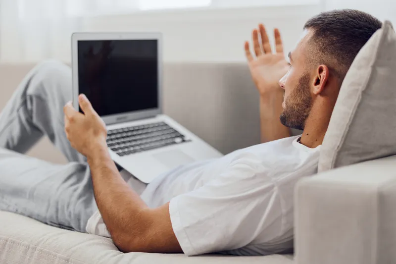 Engaged man communicating online while lounging at home, wearing casual attire and relaxed expression, enjoying a video call in a cozy setting.