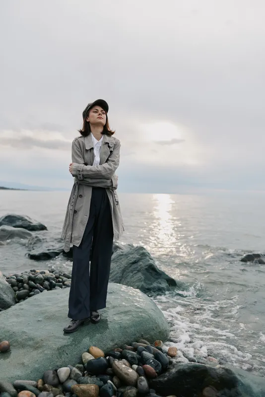 Confident woman in casual clothes and hat stands on rocky shore with arms crossed, looking at calm sea during cloudy day. Concept of solitude, nature, and peaceful contemplation.