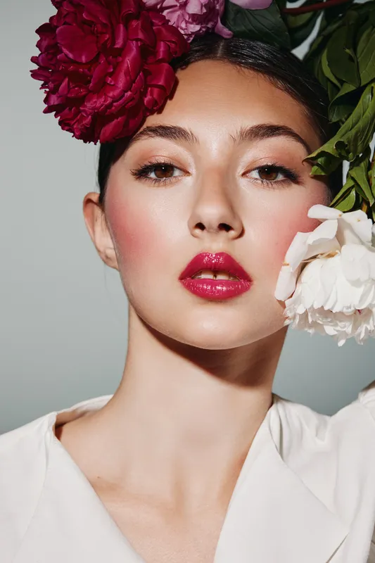 Creative beauty portrait of a young woman with glossy red lips and subtle eye makeup, adorned with vibrant red and white flowers. Studio shot with soft natural lighting. Fashion concept.