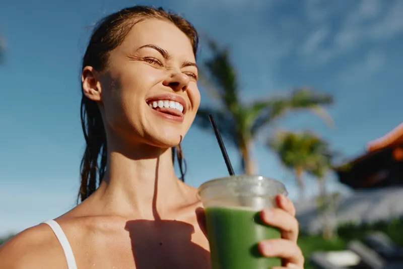 A happy woman in casualwear enjoys a green smoothie outdoors on a sunny day with palm trees in the background. Concept for healthy lifestyle and wellness.