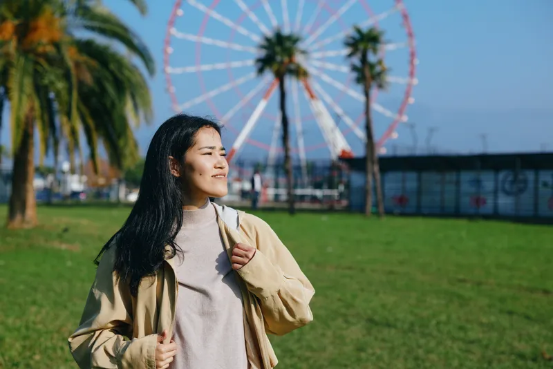 Woman smiling in a park with a ferris wheel behind, casual jacket, sunny day, enjoying outdoor leisure and travel vibes in bright scenery
