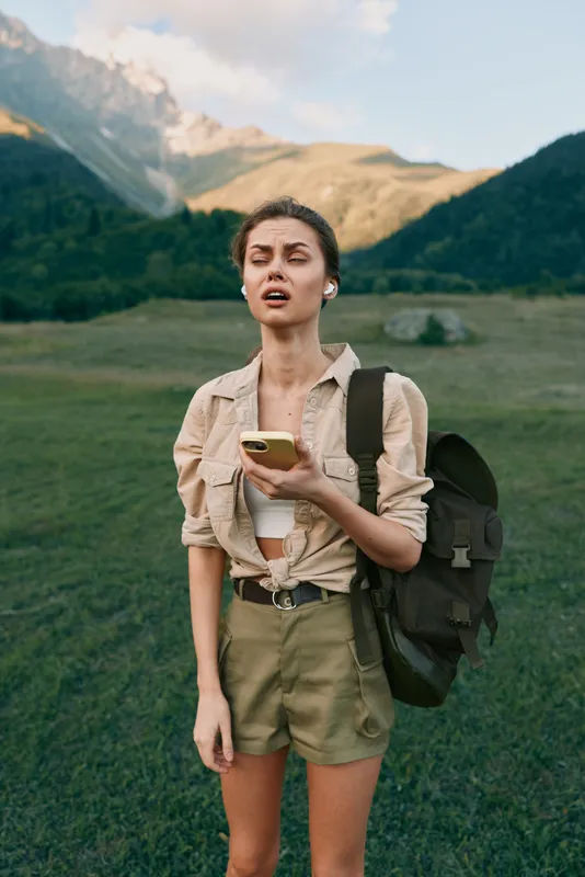 Young woman hiking in a mountain meadow holding a phone, backpack on her shoulder, wearing khaki shorts and a beige shirt.