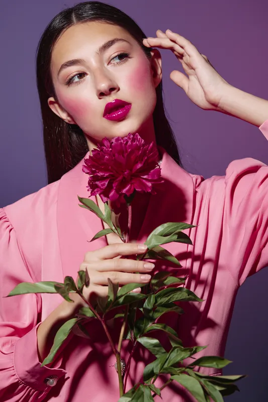 Fashion woman with glossy magenta lips and pink blush holds deep red peony. Studio portrait uses bold lighting and dramatic styling for beauty, creative, artistic fashion concept.