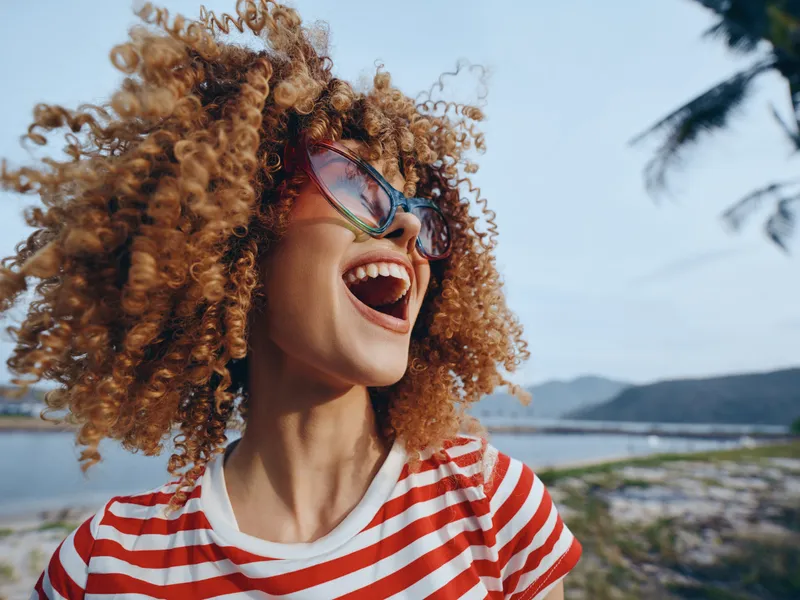 Joyful portrait of a person with curly hair and bright sunglasses laughing on a sunny beach. Vibrant energy and carefree vibe highlight playful summertime charm and expressive motion.