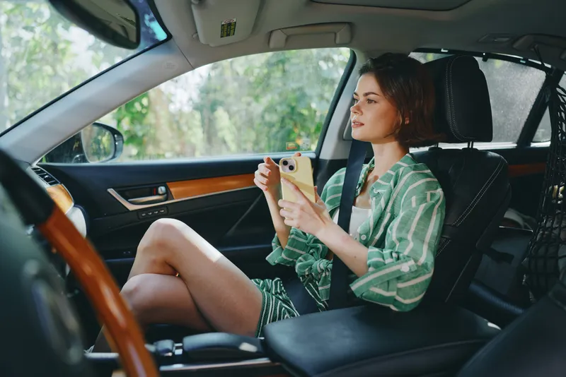 Young woman seated in car interior, casually using smartphone while parked, sunlight streaming through window.