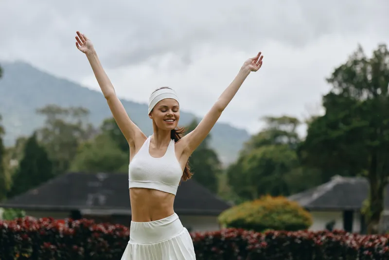 Happy young woman in sporty fashion celebrates outdoors during a workout, wearing a white crop top and skirt, with raised arms in a lush green environment