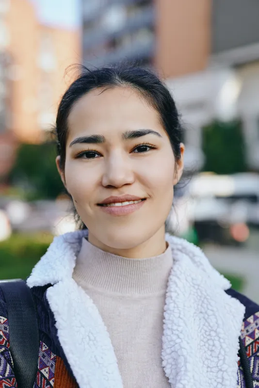 Woman with a warm smile in an outdoor urban setting wearing a fleece jacket and light sweater, daylight scene with blurred buildings in the background