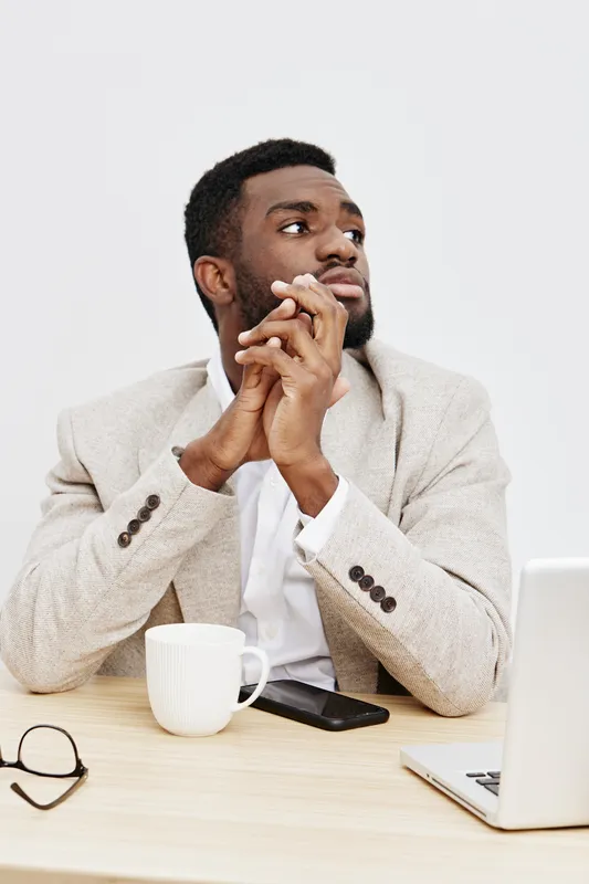 Thoughtful young man with dark skin and short black hair, wearing a beige blazer, sitting at a desk with a white mug, laptop, and glasses, looking away with a pensive expression, isolated on plain whi
