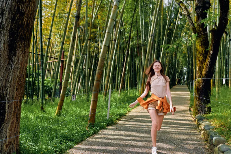 Woman walking on a gravel path through a bamboo forest, smile bright in a casual summer skirt and blouse, carefree motion with sunlight filtering through tall green bamboo.