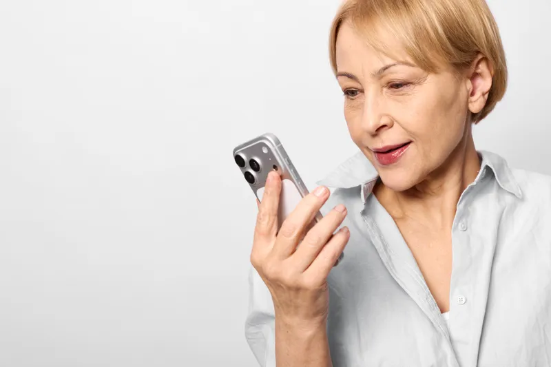 Elderly woman holds a smartphone close to her face, mobile technology communication in focus, worried concerned expression, portrait indoors on white background with copyspace