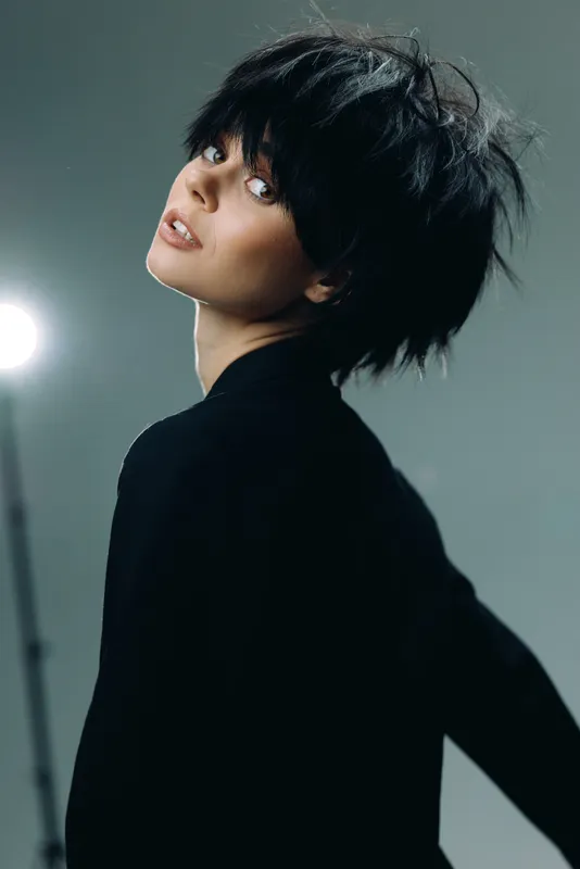 Young woman with short dark hair posing in black outfit against gray background, stylish and confident expression.