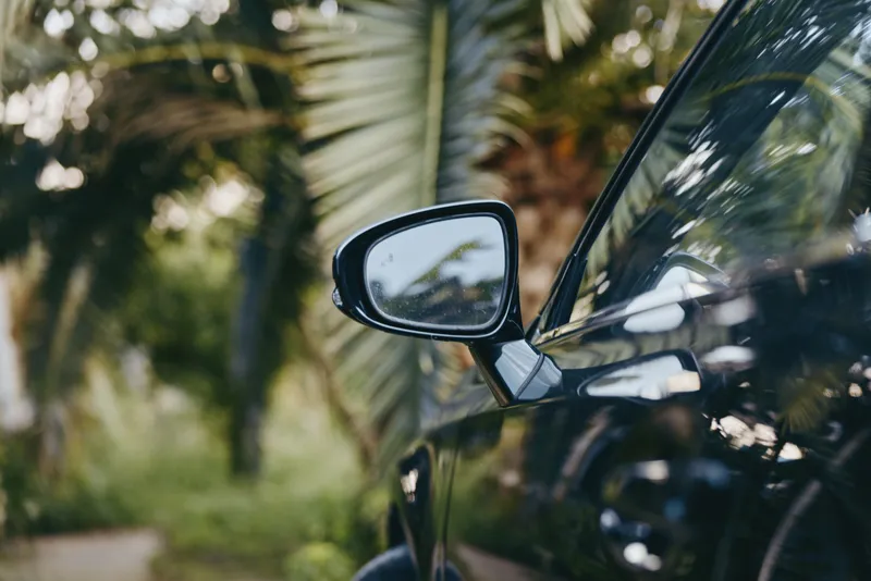 Car side mirror closeup with black car parked by palm trees in tropical setting, luxury coupe detail reflecting foliage, glossy paint and bokeh background for travel lifestyle.