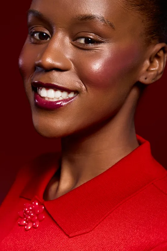 Close-up portrait of smiling black woman with dark skin wearing red blouse. She shows bright makeup and healthy skin on red background. Concept of beauty, fashion, and confidence.