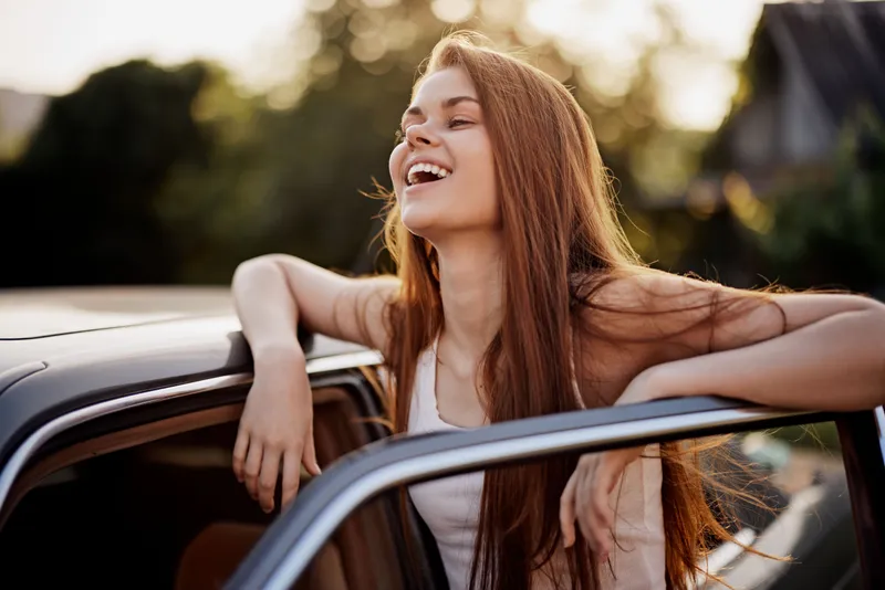Happy young woman with long hair enjoying a summer day, smiling outside a car, captured in warm sunlight with a lush green background for lifestyle photography.
