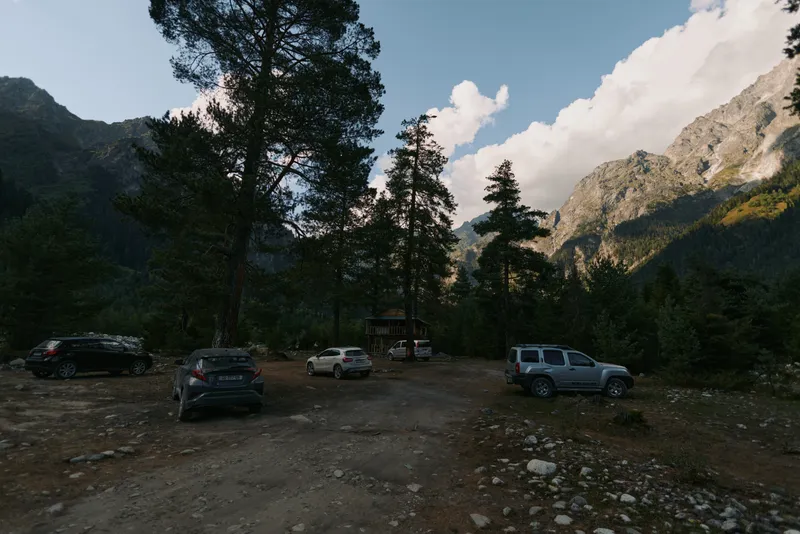 A remote mountain parking area lies among rugged terrain and tall pines. Several cars are parked while sunlight filters through clouds, creating a calm, outdoor scene.