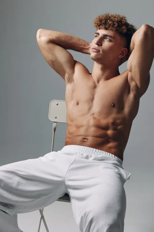 Young man sitting shirtless on chair with hands behind head showing athletic muscular torso on neutral grey background in casual white pants with relaxed confident pose in studio lighting.