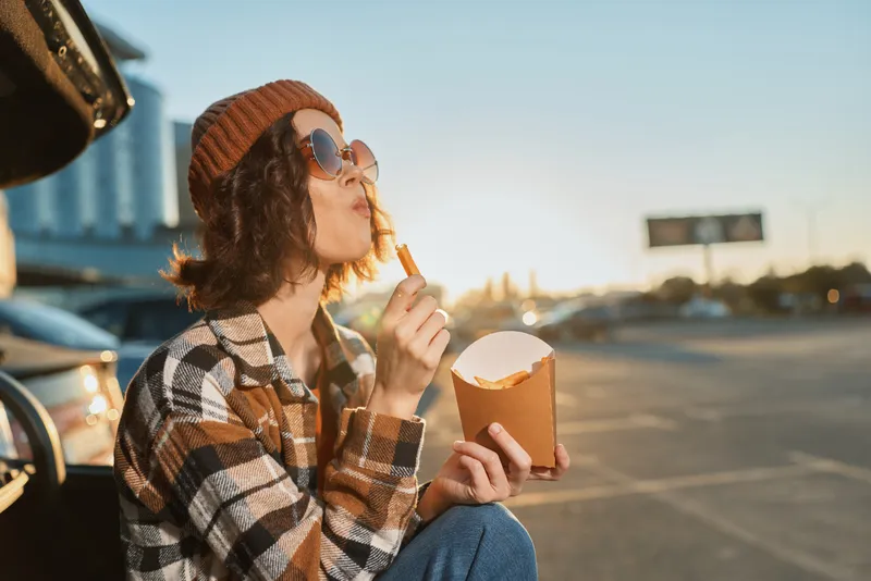 woman fries parking car sunset candid: Young woman in beanie and sunglasses eats fries from a carton by an open trunk, golden hour glow, authenticity and mindful living in an emotional storytelling