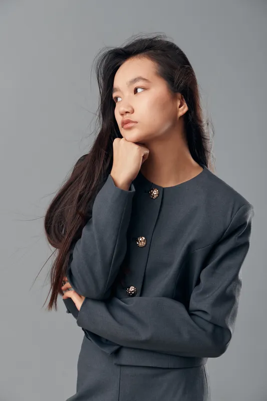 Young woman in gray suit with long black hair posing thinking and looking aside on gray background, casual fashion and elegant posture, studio portrait of confident person.