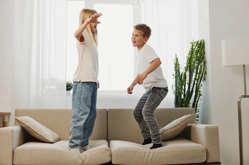 Two happy children jump and play on the sofa in a bright living room with natural light and green plants, expressing joy and energetic family moments indoors.
