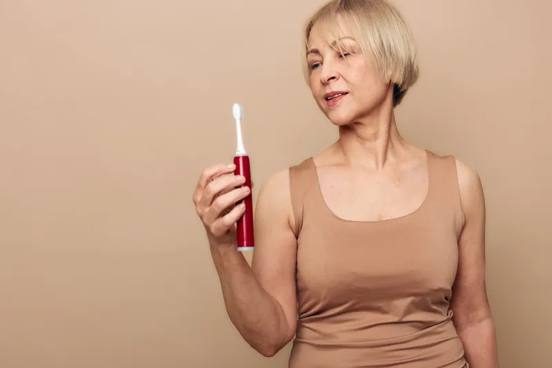 Mature woman in beige top examines red electric toothbrush with slight smile. Neutral background highlights personal care and oral hygiene concept for middle aged people.