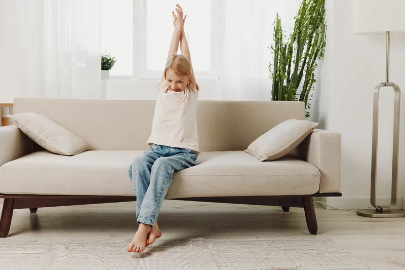 Happy little girl stretching arms while sitting barefoot on beige sofa in bright living room. Concept of morning routine, comfort, home lifestyle, and childhood relaxation.