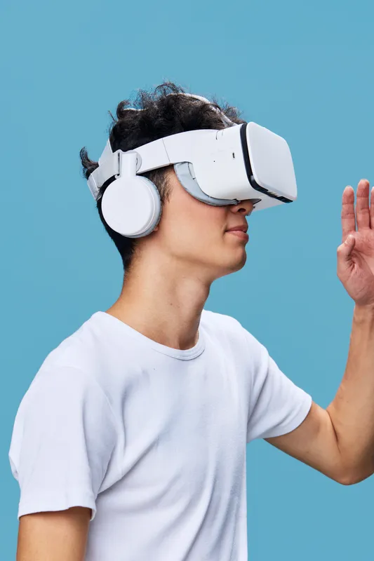 Young man wearing virtual reality headset and white headphones exploring immersive digital environment with raised hand gesture on blue background technology and innovation concept.