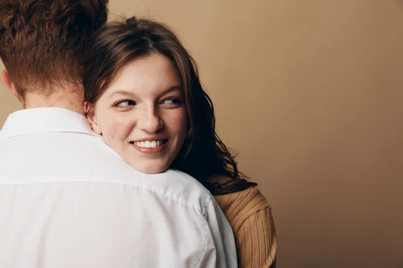 Smiling young woman with curly hair embraces man wearing white shirt, looking away with happy expression. Intimate moment, love, relationship, and affection concept on beige background.