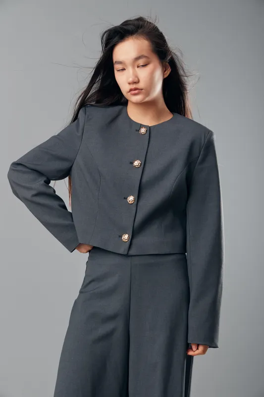 Young woman wearing dark gray pantsuit with decorative buttons posing with eyes closed against neutral background. Fashion model in elegant tailored outfit with long hair and relaxed expression.