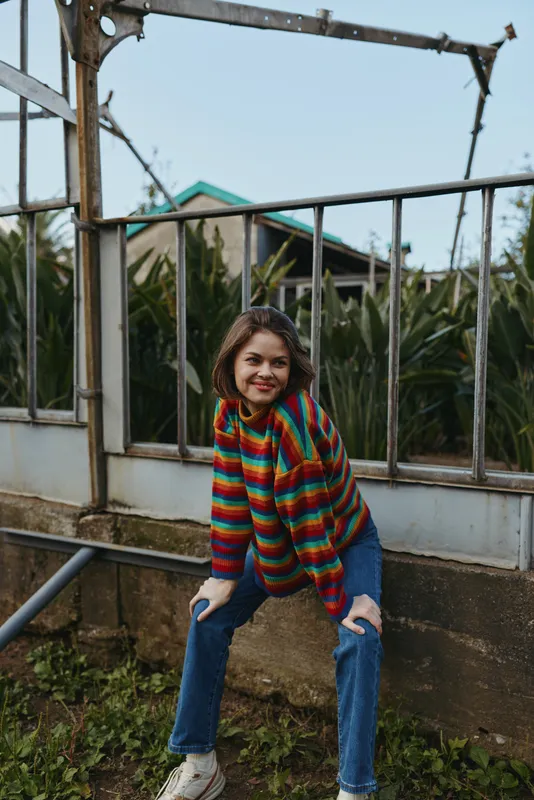 young woman, striped sweater, ripped jeans, smiling, outdoor, leaning, casual outfit, tropical plants — 25-year-old female model in a colorful rainbow knit sweater and distressed jeans sitting on a