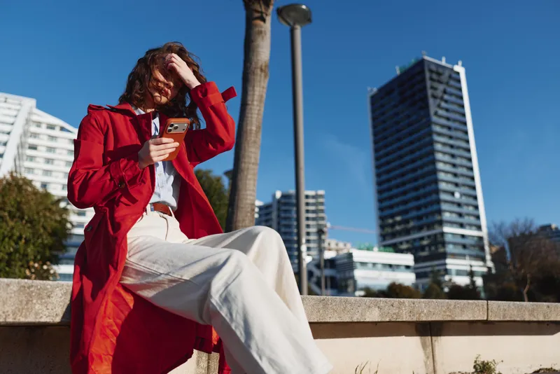 Young woman in a red trench coat sits on a concrete ledge outdoors while using her smartphone on a sunny day in the city.