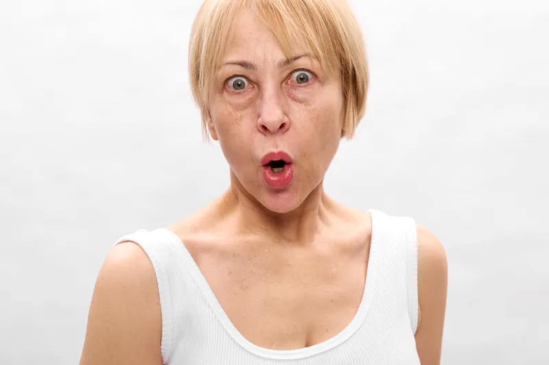 Surprised elderly woman, closeup face, open mouth expression, looking at camera, studio white background, neutral soft lighting, sleeveless casual top, startled emotion