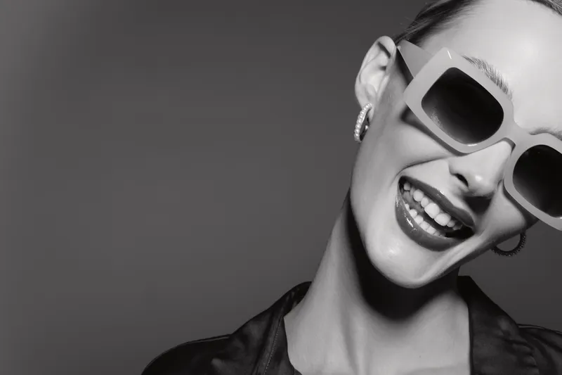 Happy Caucasian woman with pale skin wearing large sunglasses smiles broadly against gray background.