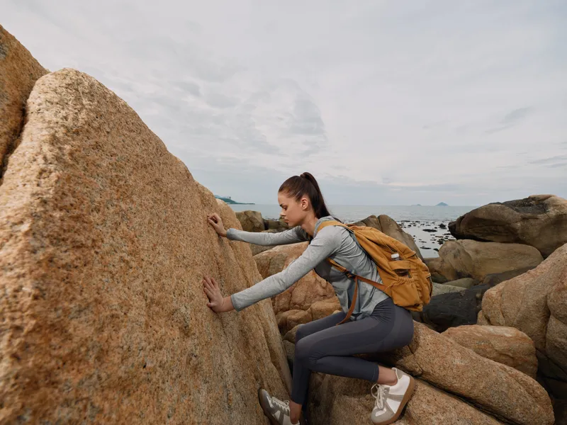 Young woman climbing on rocky coastline, showcasing determination and adventure in outdoor sportswear, set against a scenic backdrop with soft grey clouds