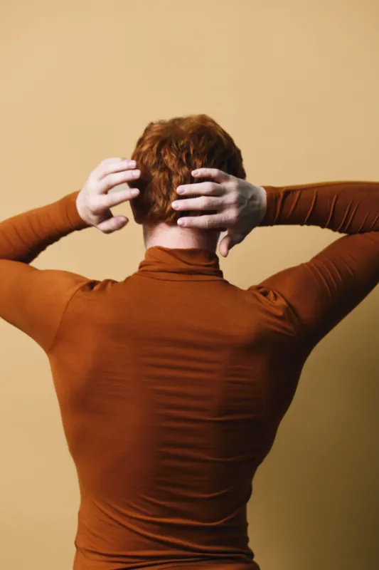 Man with red hair wearing brown turtleneck stands with hands on head against beige background in studio. Back view of male model with casual fashion and relaxed pose for portrait.