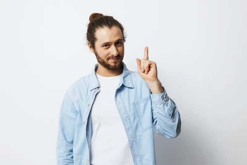 Young man with long hair wearing a light blue shirt showing a finger up, symbolizing a bright idea or success in a minimalistic background