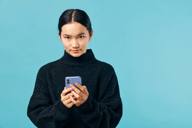 Young woman with dark hair in black sweater holding smartphone with both hands standing against blue background looking at camera with calm expression, casual and modern style portrait.