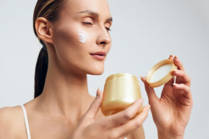 Young woman with closed eyes applying face cream on cheek, holding open jar of cream. Concept of skincare, beauty, self-care, and natural wellness in soft natural light.