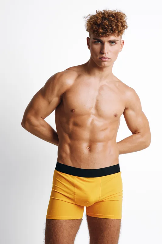 Young muscular man with curly hair poses shirtless wearing yellow underwear while standing against white background. Fit body, tanning skin and confident expression in studio lighting.