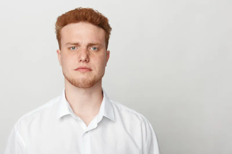 Portrait of serious young man with red hair and beard wearing white shirt standing against plain light background. Concept for confidence, professionalism, and casual style.