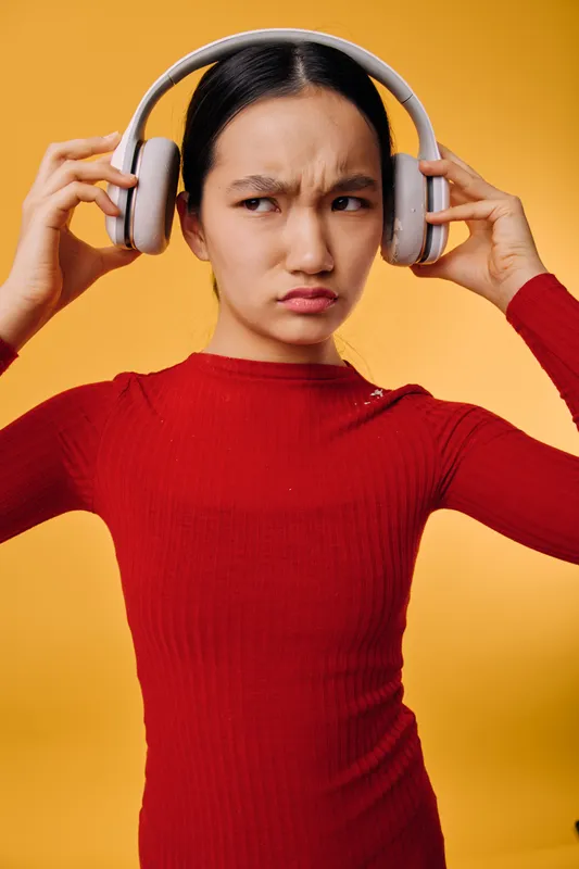 Young woman in red sweater adjusting wireless headphones with a confused expression on orange background. Girl listens to music with furrowed brows and dislike feeling in studio.
