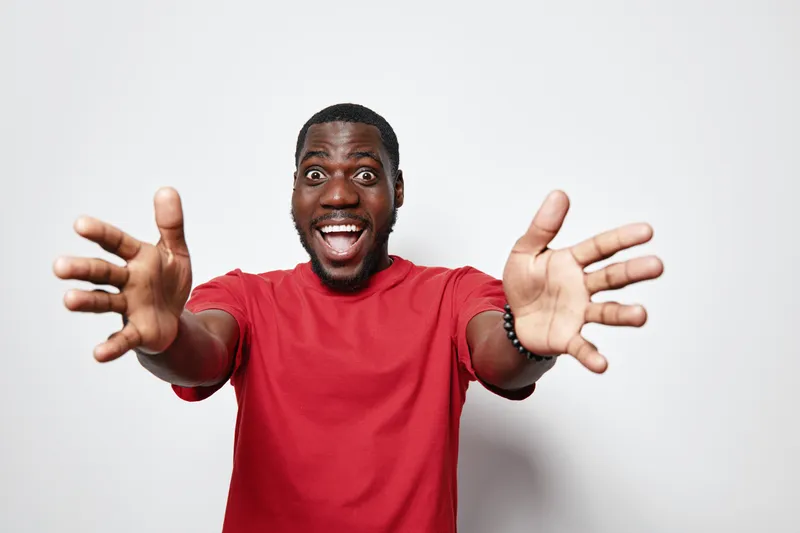 African American man with dark skin wearing red shirt smiling and reaching out with open hands for hug or welcome on white background in studio portrait.