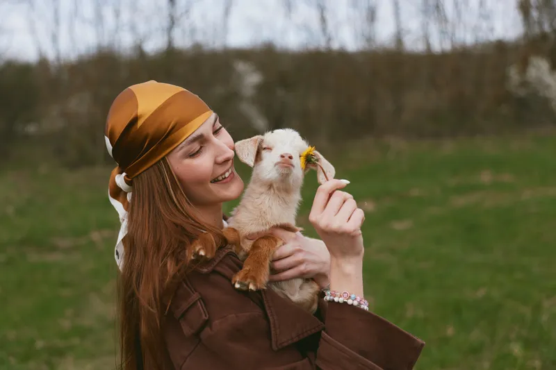 Joyful woman in a brown jacket hugs a playful goat kid outdoors, admiring a small flower held near the animal, in a rural field with soft natural light