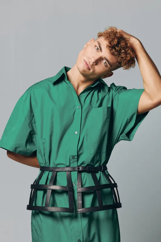 Young man with curly hair wearing green shirt dress and unique black harness belt posing against plain background in studio. Fashion model with casual distinctive style and stoic expression.
