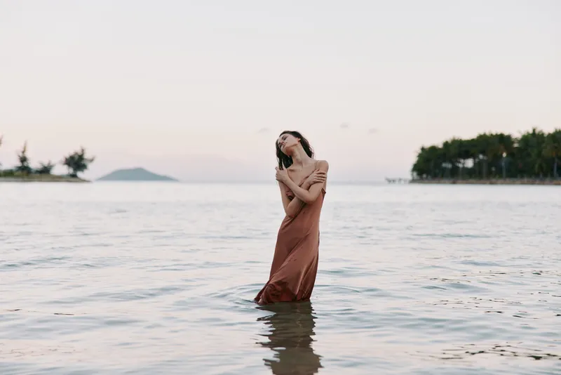 A solitary woman stands in shallow sea water at sunset, wrapped in a flowing brown dress, creating a contemplative, serene moment as gentle waves lap around her.