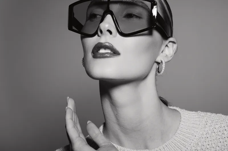 Young white woman with pale skin wearing oversized sunglasses and hoop earrings poses in black and white studio portrait.