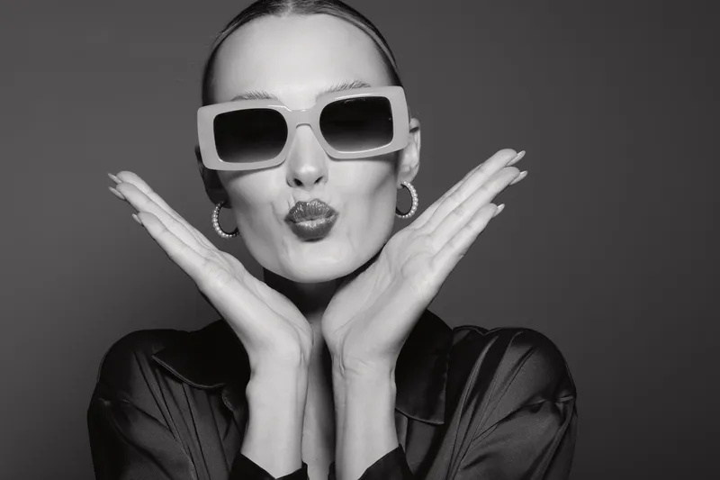 Stylish young Caucasian woman with light skin wearing oversized sunglasses and hoop earrings makes a playful kiss face with hands near cheeks in studio. Concept of fashion and fun.
