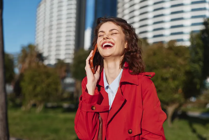 Young woman with curly hair wearing a red coat laughs joyfully while talking on the phone outdoors in a sunny urban park setting.