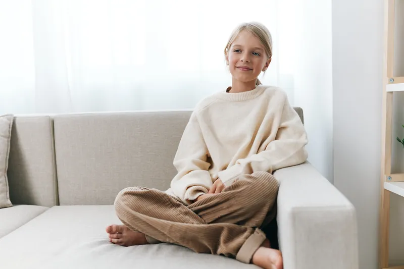 A young girl sits on a light sofa in a cozy living room, wearing a cream sweater and brown pants, smiling softly at the camera in a warm, relaxed setting.