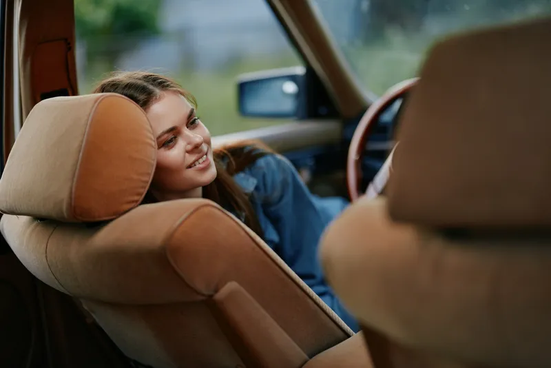 Smiling young woman relaxing in a vintage car, embodying a carefree spirit with natural light and warm tones, perfect for lifestyle and travel photography.