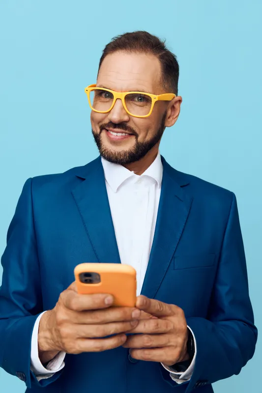 A confident business professional stands against a bright colored background, focused on a smartphone with a warm smile, illustrating a modern, energetic work moment and clear communication.